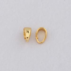 May include: Two gold-coloured pendant bails. One bail is rectangular with a curved top, and the other is oval-shaped. Both are made of shiny, polished metal, likely for jewellery making or crafting.