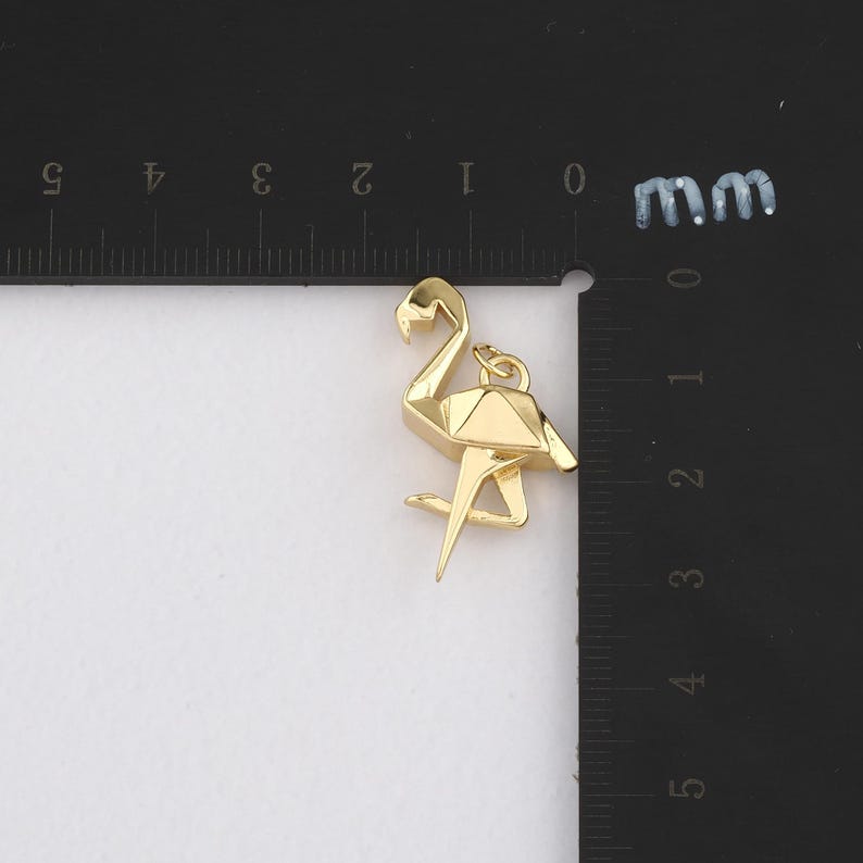K&ouml;nnte beinhalten: Ein goldfarbener Flamingo-Anh&auml;nger mit geometrischem Design. Der Anh&auml;nger ist etwa 2,5 cm hoch und hat eine Schlaufe zum Befestigen an einer Kette oder einem Armband. Der Flamingo steht auf einem Bein.