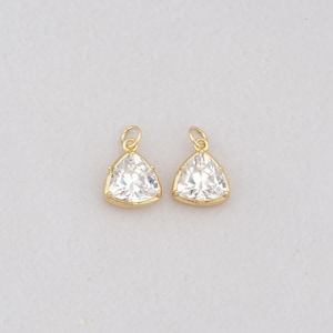 Puede incluir: Dos dijes de joyería en tono dorado, cada uno con una piedra preciosa transparente de corte triangular facetado engastada en un marco de metal con una pequeña argolla circular en la parte superior para colgar. Las piedras son claras.