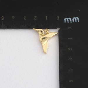 K&ouml;nnte beinhalten: Ein goldfarbener Kolibri-Anh&auml;nger. Der Anh&auml;nger ist etwa 2,5 cm lang und hat oben eine Schlaufe zum Befestigen an einer Kette. Der Kolibri ist in einem stilisierten Design aus gefaltetem Papier dargestellt.