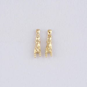 May include: Two gold-coloured pendants shaped like hands making the 'I love you' sign. Each pendant has a loop at the top for a chain. The pendants are displayed against a white background.