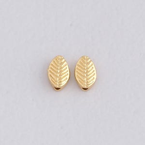 May include: Two gold-colored leaf-shaped beads. Each bead has a detailed leaf vein pattern and a small hole at the bottom. The beads are oval and have a shiny finish, perfect for jewelry making.