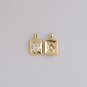May include: Two gold-colored pendants with a square frame and an oval, clear stone in the center. Each pendant has a small loop at the top for a chain. The pendants are displayed on a light gray background.