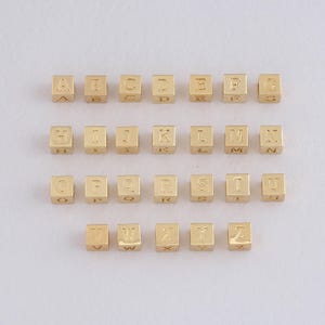 Könnte beinhalten: Goldfarbene Alphabet-Buchstabenperlen, jede würfelförmig mit einem einzelnen Buchstaben auf einer Seite graviert. Die Buchstaben A bis Z sind abgebildet, ideal für die Schmuckherstellung oder Bastelprojekte. Die Perlen sind in Reihen angeordnet.