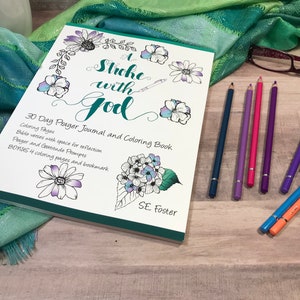 May include: A prayer journal and colouring book titled "A Stroke with God" with floral illustrations. The book includes colouring pages, Bible verses, and prompts. Coloured pencils in shades of purple, blue, and pink are arranged nearby.