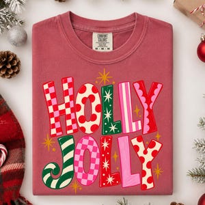 May include: A dusty rose crewneck t-shirt with the text HOLLY JOLLY printed in large, multi-colored, patterned letters. The letters feature red and pink checkerboard, red polka dots, green stripes, and solid pink designs with small yellow stars.