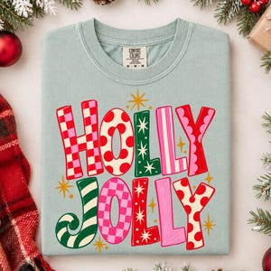 May include: A light blue crewneck t-shirt featuring the text HOLLY JOLLY in large, multi-colored letters with various patterns including red and pink checkers, red polka dots, and green stripes, accented by small yellow starburst shapes.