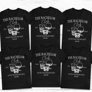 May include: Six black short-sleeve t-shirts featuring a white graphic of three glasses and the text THE BACHELOR Club EST. 2024 DAVID'S BACHELOR PARTY MIAMI. The design includes a tall glass, a short tumbler, and a rounded brandy snifter glass.