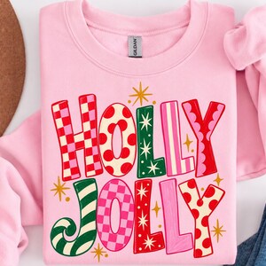 May include: A light pink crewneck sweatshirt featuring the text HOLLY JOLLY in large, multi-colored letters. The letters display various patterns including red and pink checkers, red polka dots, green with white stars, and pink stripes.
