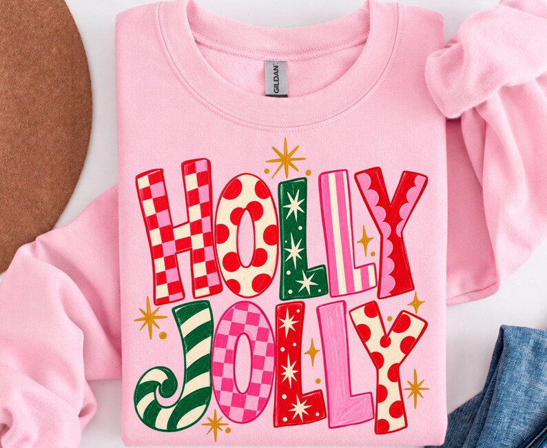 May include: A light pink crewneck sweatshirt featuring the text HOLLY JOLLY in large, multi-colored letters. The letters display various patterns including red and pink checkers, red polka dots, green with white stars, and pink stripes.