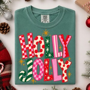 May include: A sage green t-shirt featuring the text HOLLY JOLLY in large, multi-colored, patterned letters. The letters include red and white checkerboard, red polka dots, green with white stars, and pink stripes. Small yellow star accents surround the text.