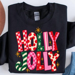 May include: A black long-sleeve sweatshirt featuring the text HOLLY JOLLY in large, multi-colored letters. The letters display various patterns including red and pink checkers, red polka dots, green stripes, and solid pink, accented by gold stars.