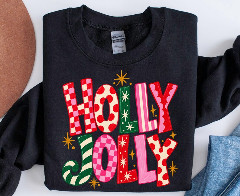 May include: A black long-sleeve sweatshirt featuring the text HOLLY JOLLY in large, multi-colored letters. The letters display various patterns including red and pink checkers, red polka dots, green stripes, and solid pink, accented by gold stars.