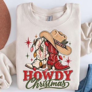 May include: A cream-colored crewneck sweatshirt featuring a graphic of a tan cowboy hat and two cowboy boots, one white and one red, wrapped in colorful Christmas lights. The text HOWDY Christmas is printed in red and green below the boots.