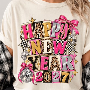 May include: A cream-colored t-shirt featuring a graphic print that reads HAPPY NEW YEAR 2027. The text is styled in block letters with leopard print, hot pink, gold, and silver textures, accented by disco balls, pink bows, and star shapes.