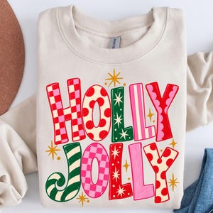 May include: A sand-colored crewneck sweatshirt featuring the text HOLLY JOLLY in large, multi-colored letters. The letters display various patterns including red and pink checkers, red polka dots, green stripes, and solid pink and red colors.