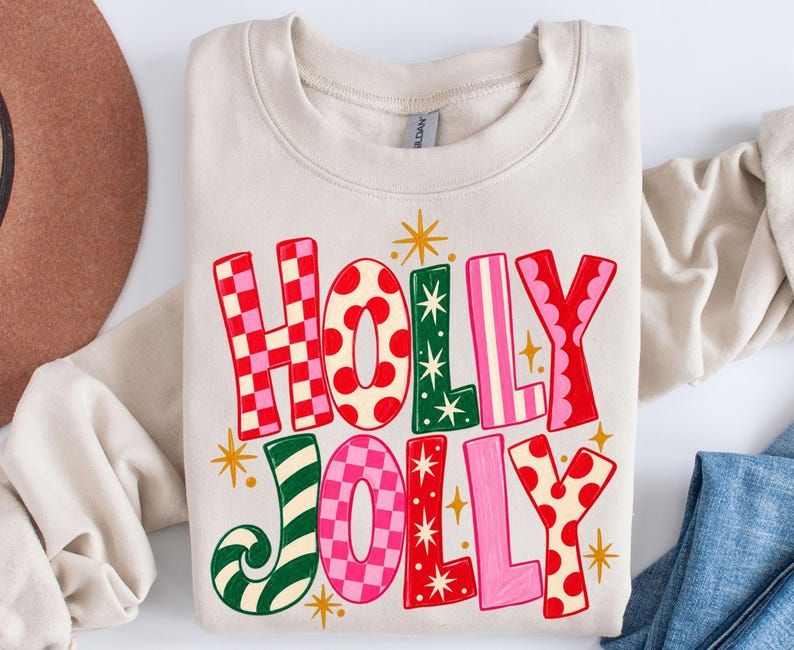 May include: A sand-colored crewneck sweatshirt featuring the text HOLLY JOLLY in large, multi-colored letters. The letters display various patterns including red and pink checkers, red polka dots, green stripes, and solid pink and red colors.