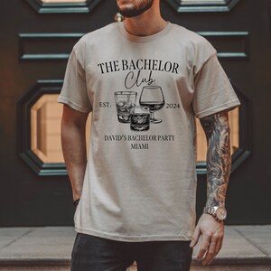 May include: A beige short-sleeve t-shirt featuring black text that reads THE BACHELOR Club EST. 2024 DAVID'S BACHELOR PARTY MIAMI. The graphic includes illustrations of three different types of glassware filled with liquid and ice cubes.