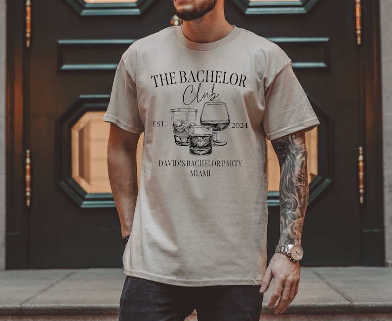 May include: A beige short-sleeve t-shirt featuring black text that reads THE BACHELOR Club EST. 2024 DAVID'S BACHELOR PARTY MIAMI. The graphic includes illustrations of three different types of glassware filled with liquid and ice cubes.