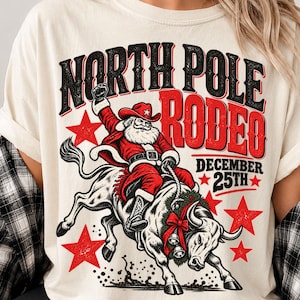 May include: A cream-colored t-shirt featuring a graphic of Santa Claus riding a bucking bull. The text reads NORTH POLE RODEO DECEMBER 25TH. The design includes red stars, a bull wearing a wreath with bells, and Santa in a cowboy hat.