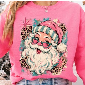 May include: A bright pink sweatshirt featuring a graphic print of a winking Santa Claus wearing teal-rimmed glasses and a matching hat. The design includes leopard print accents, teal brush strokes, and pink starbursts on a soft fabric base.