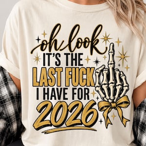 May include: A cream-colored t-shirt featuring a graphic print with the text "oh look IT'S THE LAST FUCK I HAVE FOR 2026" in black and gold lettering. The design includes a skeletal hand making a middle finger gesture adorned with a gold ribbon bow.