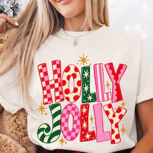Holly Jolly Christmas shirt with oversized hand-drawn red, pink, green and cream lettering featuring checkerboard, polka dot and striped patterns with retro gold starbursts, shown on adult Comfort Colors tees and Gildan sweatshirts.