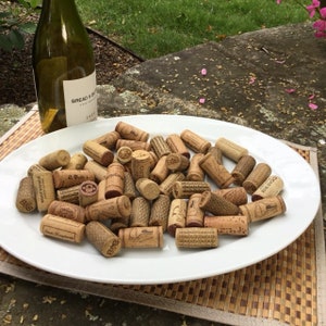 May include: A white ceramic plate filled with a collection of used wine corks. The corks are various shades of brown and have different textures and markings. A bottle of wine is visible in the background.