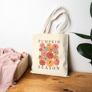 Peut inclure: Un sac fourre-tout en toile couleur crème doté de deux longues anses et d'une impression graphique centrale. Le motif présente une collection de citrouilles stylisées dans des tons orange, rouge et vert sourd, avec le texte PUMPKIN SEASON imprimé.