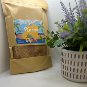May include: A brown paper bag with a label that says "Tropical's Kasav" and a picture of cassava root. The bag is filled with a brown, shredded product. The bag is sitting on a white surface next to a potted plant with purple flowers.