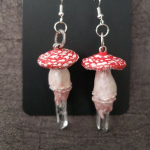 May include: Handmade earrings featuring a red and white mushroom with a clear quartz crystal stem. The earrings are on a black card with the word "Handmade" written on it.