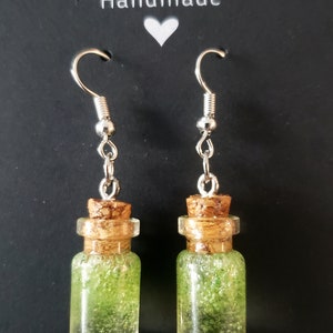 May include: A pair of silver dangle earrings with green glitter filled glass bottles and cork stoppers. The earrings are labelled "Handmade".