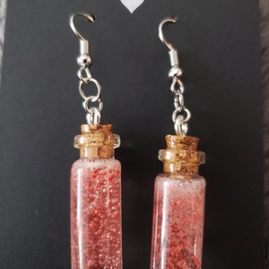 May include: A pair of handmade earrings featuring small glass bottles filled with a pink liquid and glitter. The bottles have cork stoppers and are attached to silver hooks.