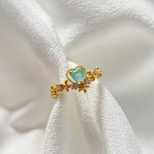 May include: A gold ring featuring a light blue heart-shaped stone, surrounded by small white stones and gold floral accents.