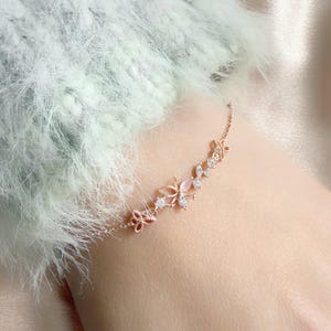 Peut inclure: Un bracelet délicat en or rose avec un motif floral orné de papillons et de feuilles. Le bracelet comprend de petites pierres précieuses transparentes et une pierre précieuse rose. Le bracelet est porté au poignet.