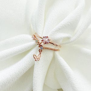 May include: A set of three delicate rose gold rings featuring a pink gemstone and a dangling butterfly charm. The rings are adorned with small, colorful crystals.