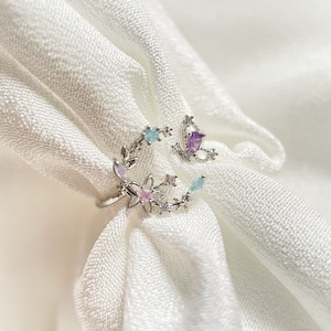 May include: A silver ring featuring a crescent moon design embellished with pastel pink, blue, and purple gemstones. The ring is detailed with delicate floral accents and a small butterfly charm.