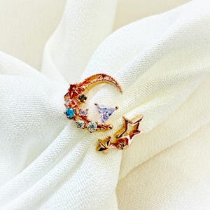 May include: A gold-coloured ring featuring a crescent moon design adorned with colourful gemstones and a star accent. The ring has a delicate, open design with a triangular purple stone in the centre of the moon.
