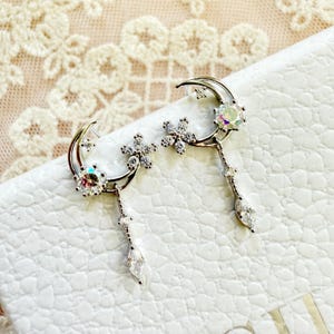 Peut inclure: Boucles d'oreilles argentées avec un croissant de lune, une pierre précieuse irisée et un cristal en forme de goutte suspendu. Les boucles d'oreilles comportent également un motif floral et de petites étoiles, créant un thème céleste.