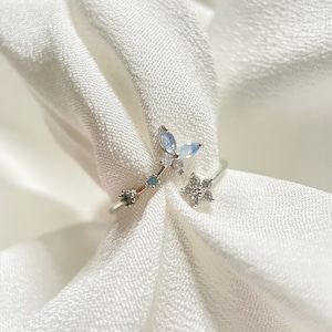 May include: A silver ring with a delicate butterfly design featuring blue and white gemstones. The ring also has a small flower design with white gemstones.