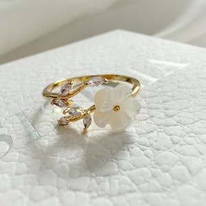 May include: Gold ring featuring a white flower and clear crystal accents. The flower is made from mother of pearl.