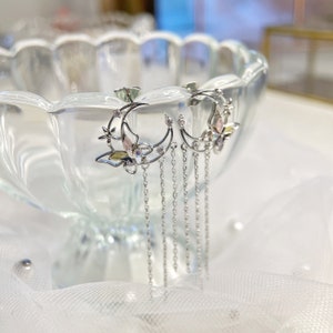 May include: Silver crescent moon earrings with a butterfly charm and dangling chains. The butterfly has colourful enamel details and clear rhinestones.