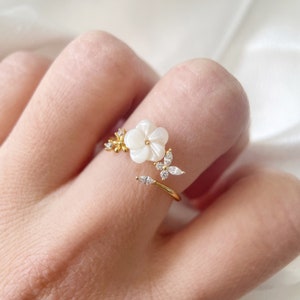 May include: A gold ring featuring a white flower and butterfly design. The flower is crafted from mother of pearl and the butterfly is adorned with clear rhinestones.