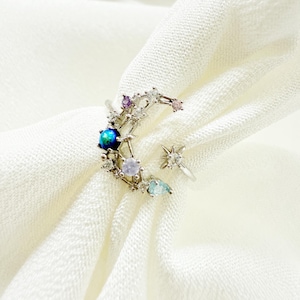 May include: A silver ring with a crescent moon design, featuring a blue and green gemstone, surrounded by smaller white, pink, and blue gemstones.