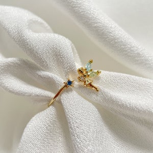 May include: Gold ring with a fairy design featuring blue and green gemstones. The fairy is holding a star with a blue gemstone.