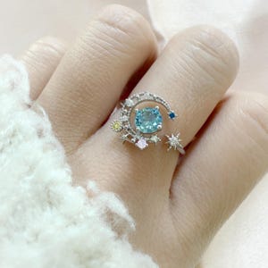 May include: A silver ring featuring a large, round, light blue gemstone set within a crescent moon design. The ring is adorned with small stars and colorful accent stones, including yellow, pink, and blue. The ring is on a finger.