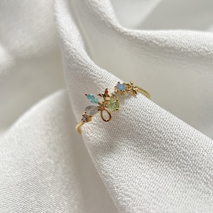 May include: A gold ring with a delicate design featuring a cluster of colorful gemstones. The ring is set on a white fabric background.