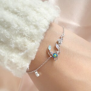 May include: A delicate silver bracelet featuring a celestial design. The bracelet has a chain with a small, sparkling charm. The main design includes a blue and gold planet-like center, surrounded by clear and blue crystals, and a crescent moon shape.