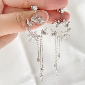 May include: Elegant silver earrings featuring a floral wreath design with dangling chains. The earrings are adorned with clear, sparkling gemstones and leaf accents, creating a delicate and sophisticated look. The earrings are designed to be worn with pierced ears.