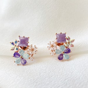May include: A pair of rose gold earrings with a cluster of purple, blue, and white flowers. The earrings feature a square purple gemstone in the center of each flower cluster.